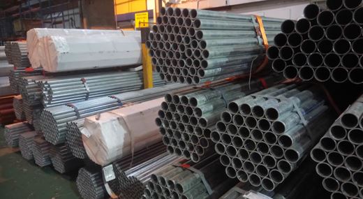 Stacks of metal pipes bundled together and stored in a warehouse. The pipes are cylindrical and appear to be made of steel or a similar metal. Some bundles are wrapped in protective coverings, and the pipes are organized in a neat and orderly manner.